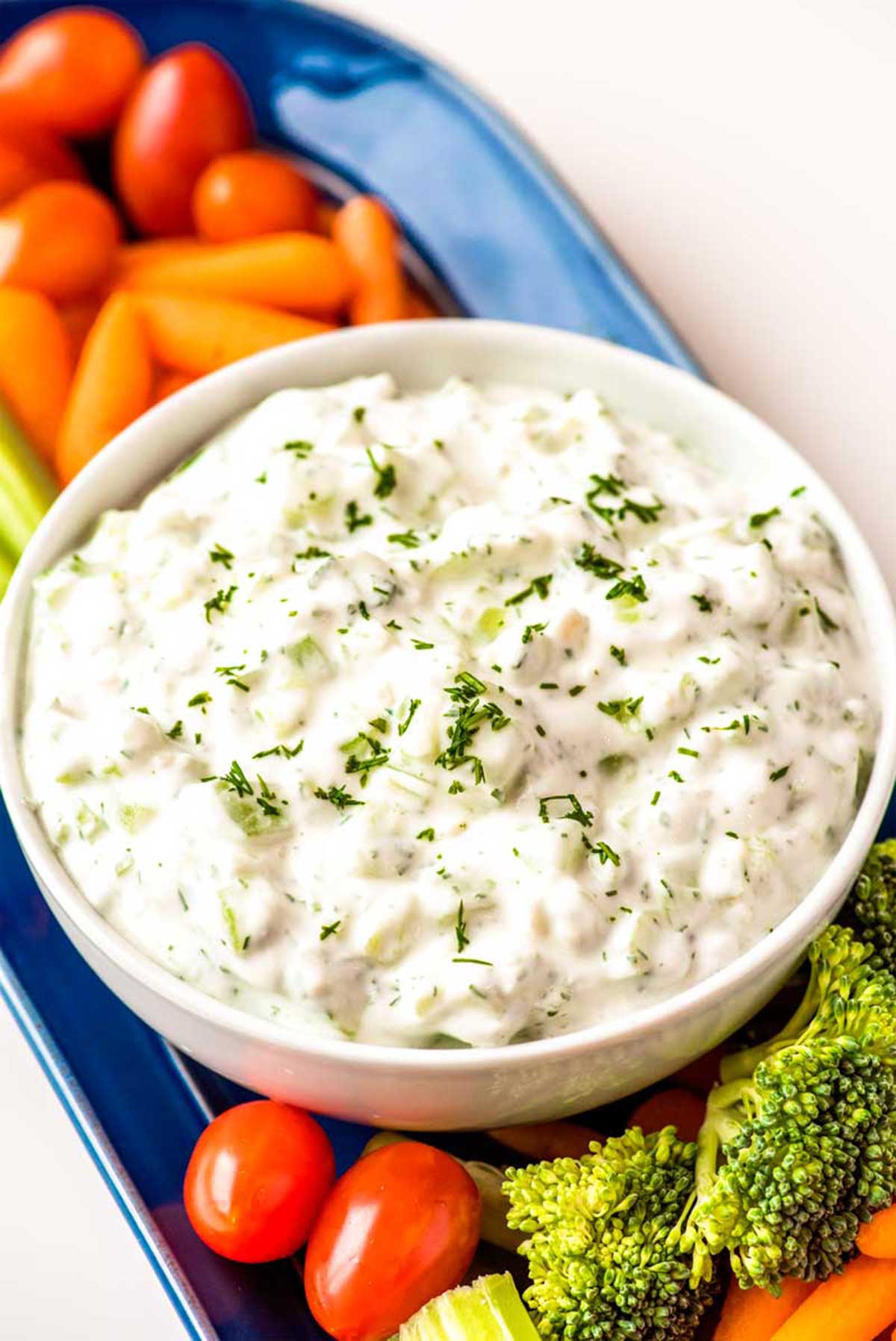 Top down view of a bowl of tzatziki sauce on a narrow serving plate filled with carrots, broccoli, and cherry tomatoes for dipping.