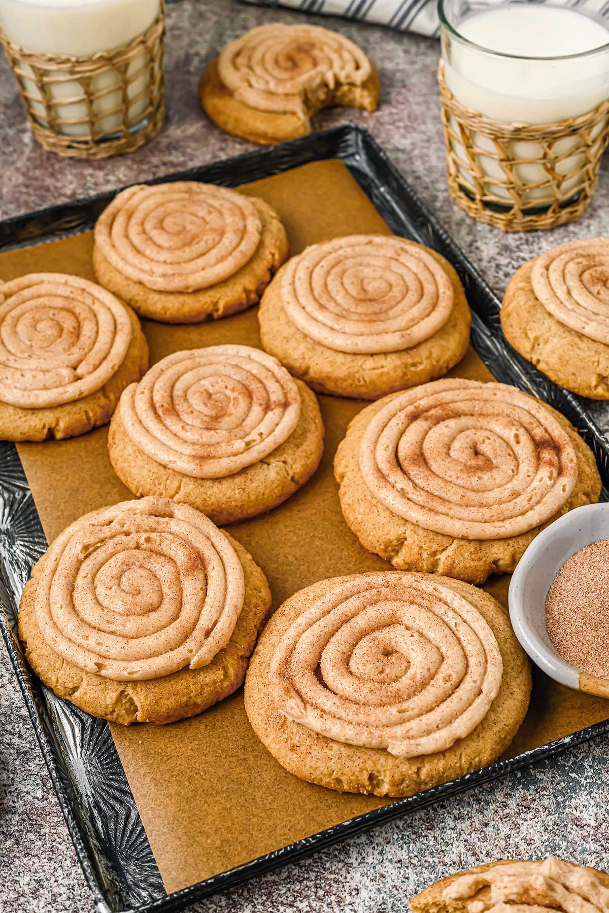 Side view of a baking sheet covered with large decorated churro cookies, pictured with glasses of milk and bowls of spices around.