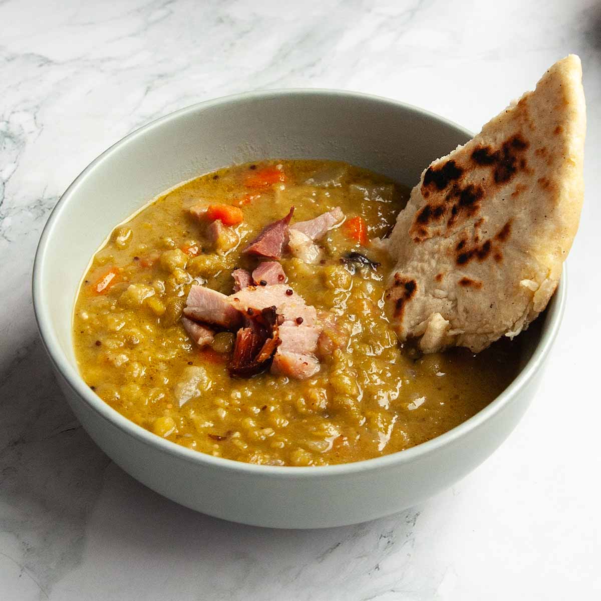 A bowl of split pea soup with ham with a piece of flatbread inside.