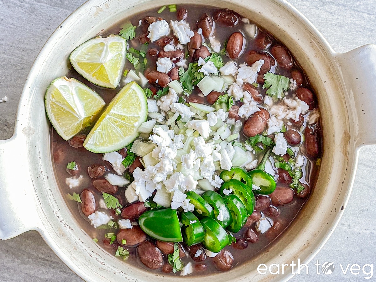 A Dutch oven full of Mexican-style beans topped with cilantro, jalapeno slices, cheese, and sliced lime wedges.
