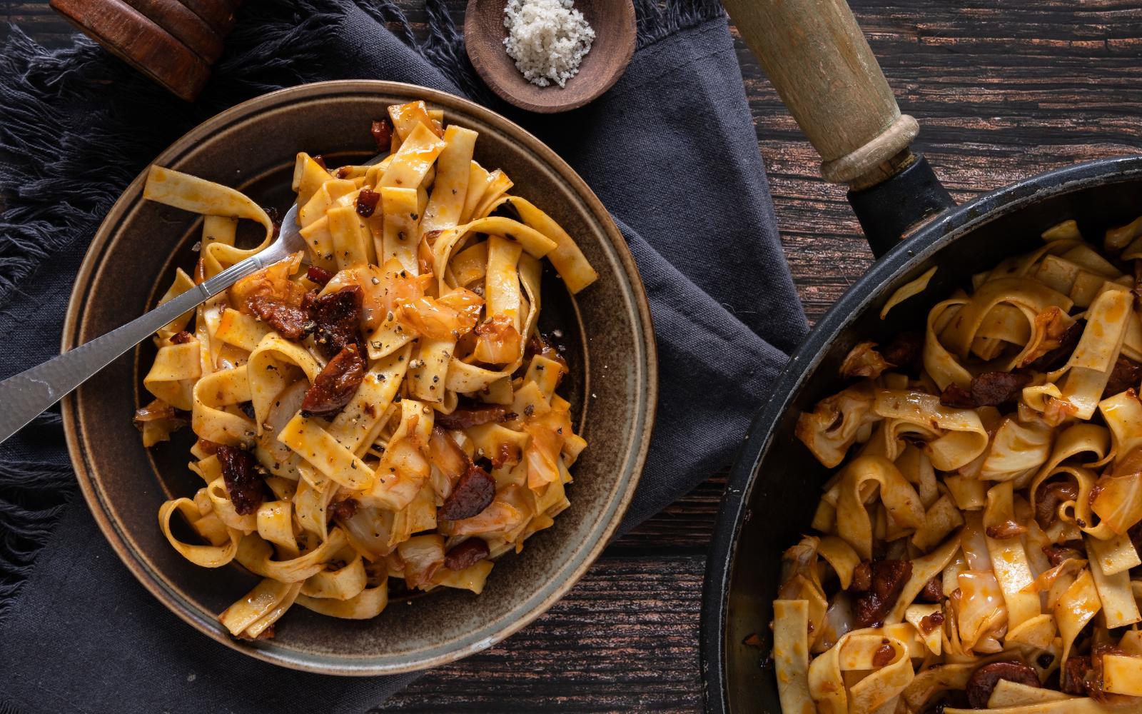 A brown bowl full of haluski, egg noodles with bacon and onion and cabbage. Slices of kielbasa are on top.