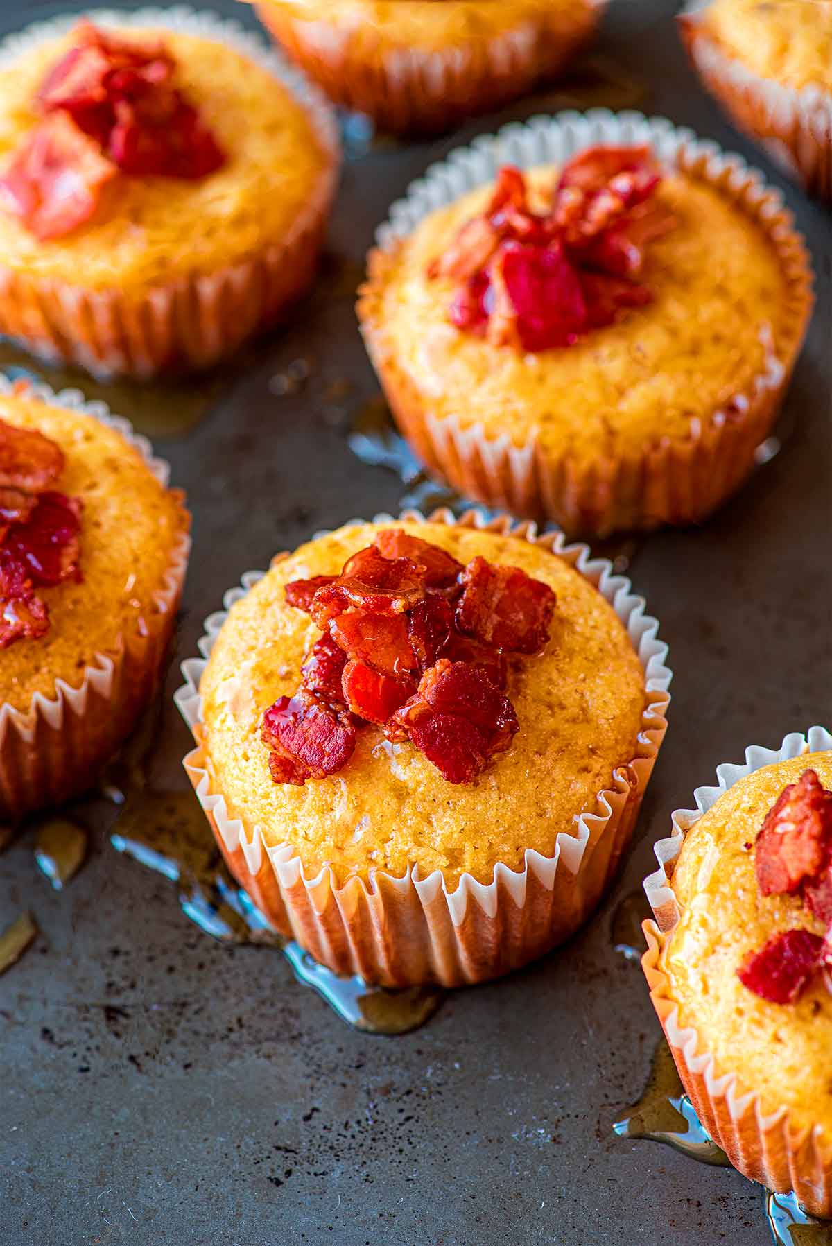 Side view of multiple prepared maple bacon cornbread muffins lined up on a baking sheet, topped with pieces of bacon and drizzled with maple syrup.