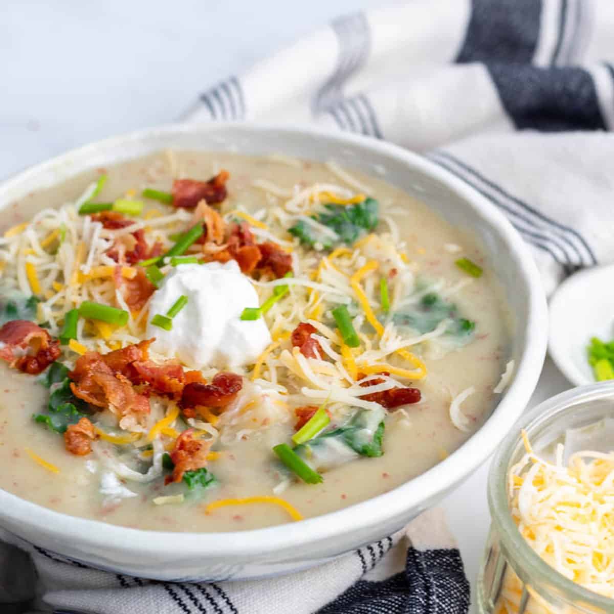 Bowl of potato leek kale soup on a blue and white napkin with bowls of shredded cheese and chives on the side.