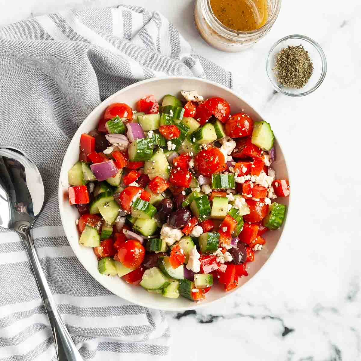 A bowl of greek salad with dressing and oregano on the side.