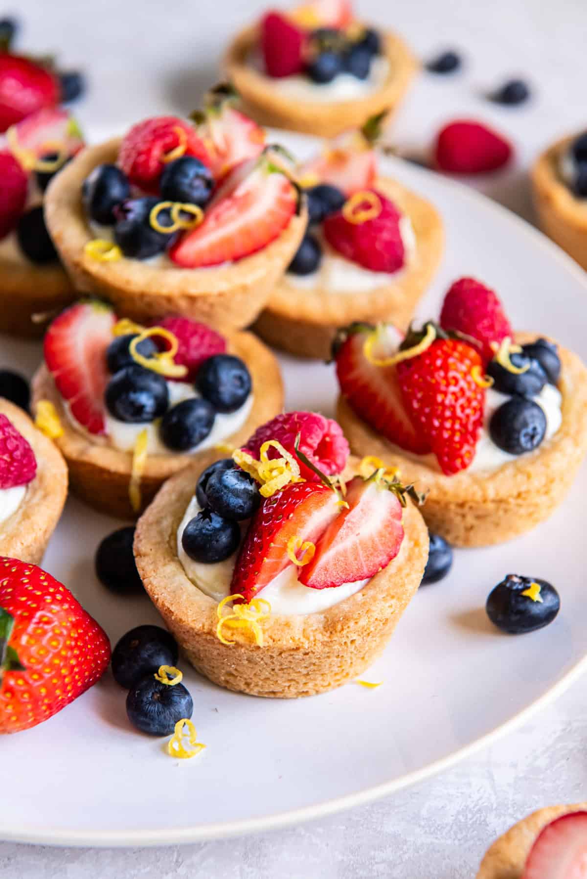 Close-up of mini fruit tartlets with cookie crusts, filled with cream and topped with fresh strawberries, blueberries, raspberries, and lemon zest on a white plate