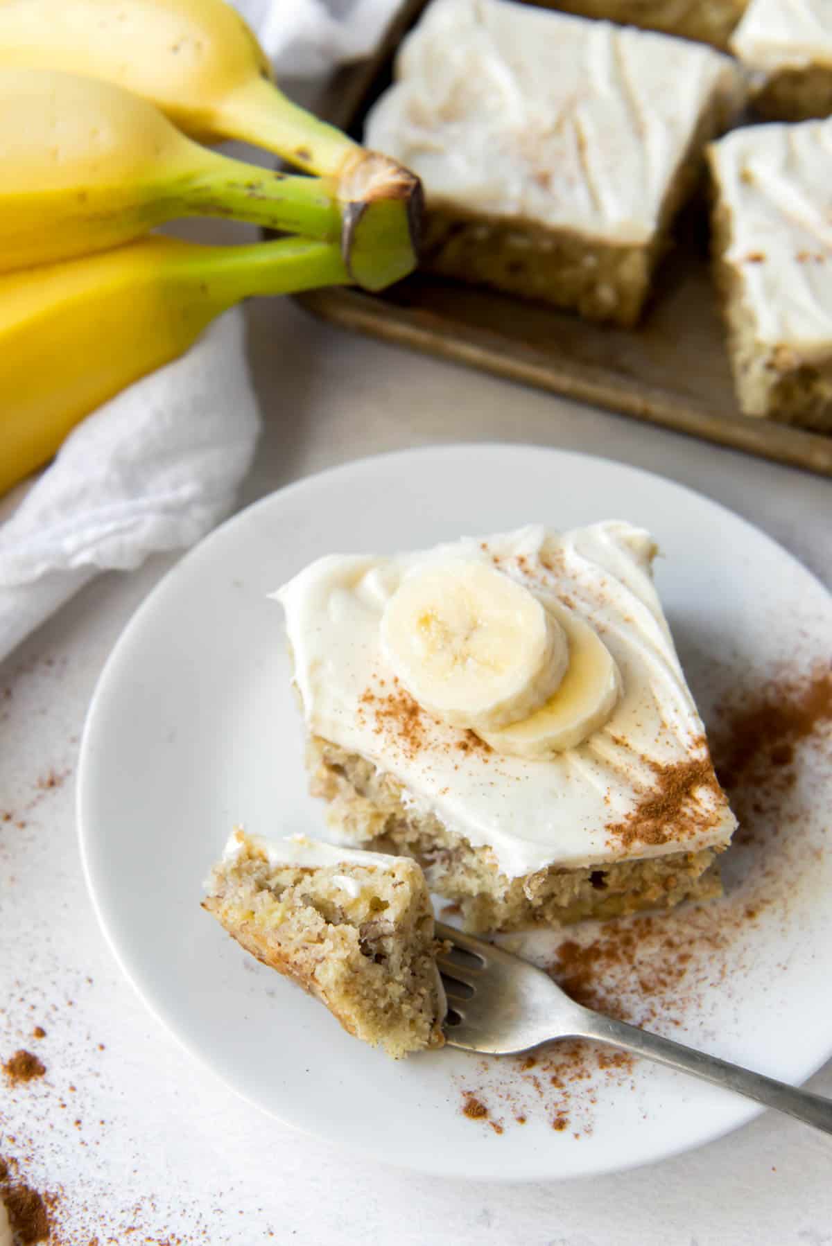 A banana bar with cream cheese icing on a plate, with bananas and more bars in the background.